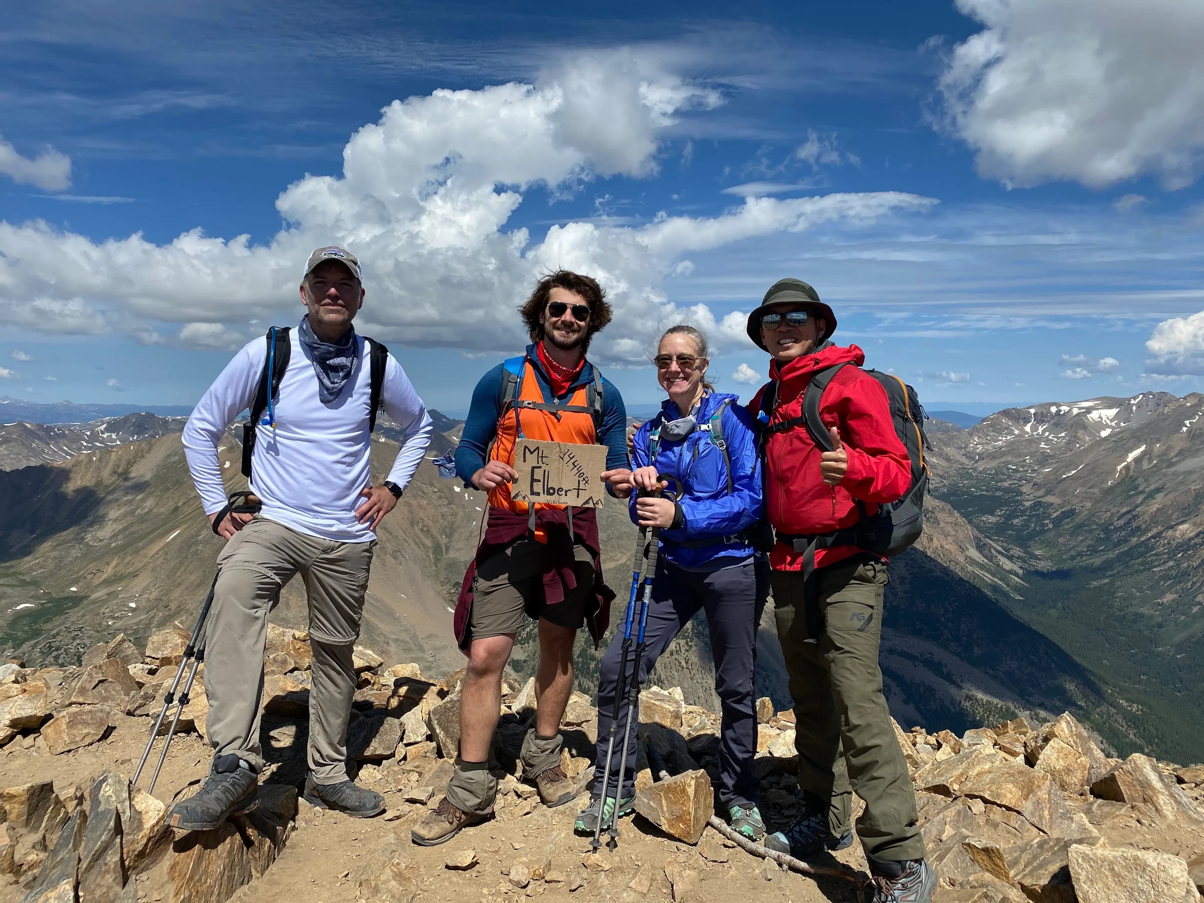 Four hikers posing on the summit of Mount Elbert with trekking gear, surrounded by a panoramic view of mountains under a bright blue sky with scattered clouds.