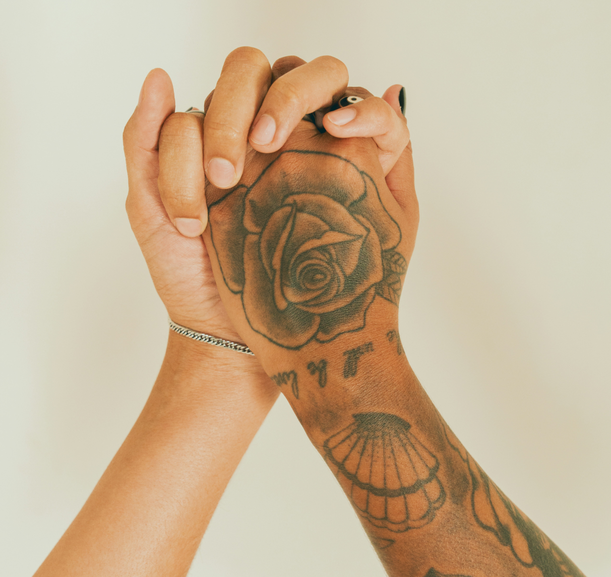 Two hands clasped together, one with intricate tattoos of a rose and seashell, symbolizing connection and diversity against a soft beige background.