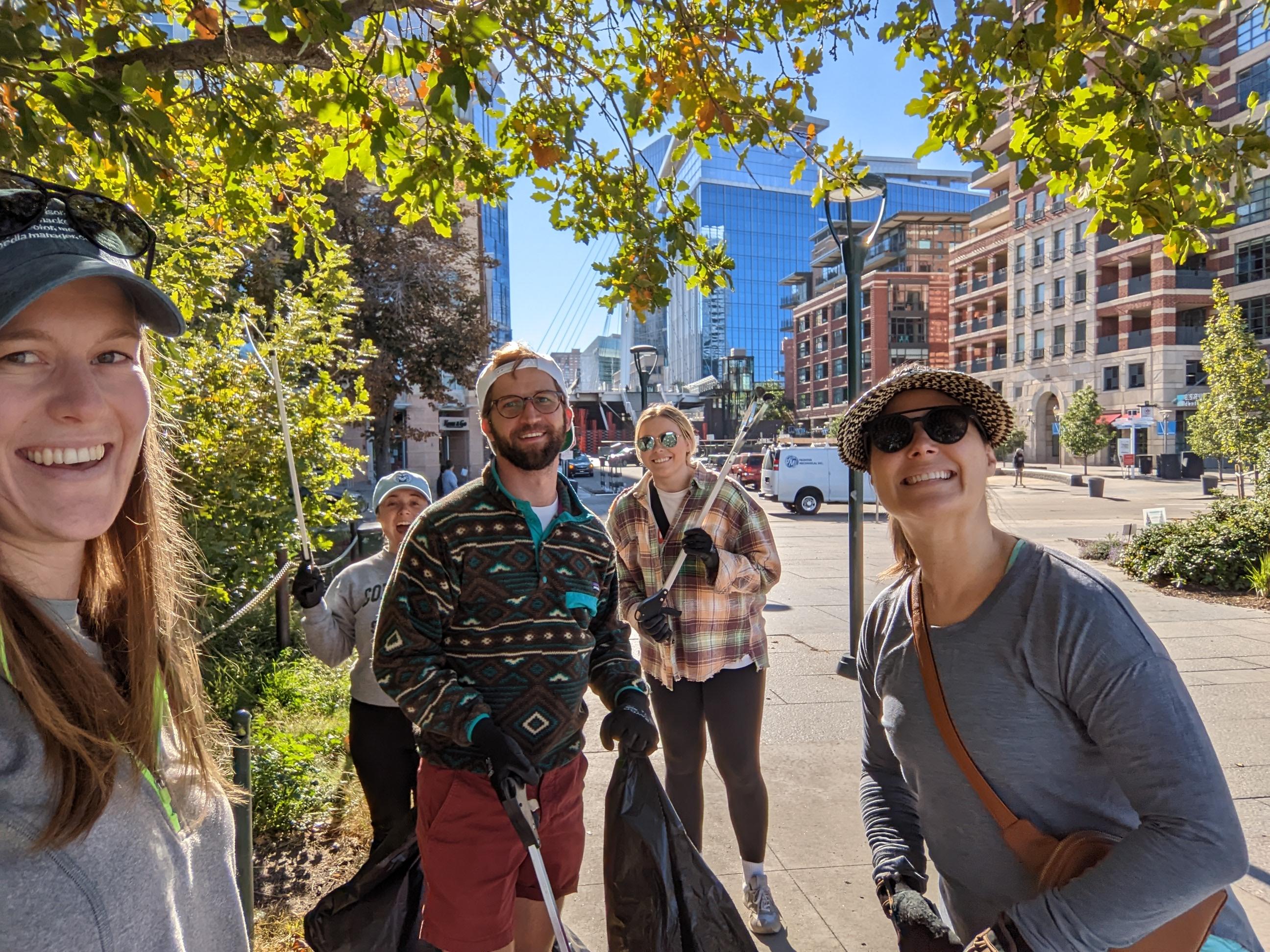 A group of smiling people in an urban area, surrounded by trees and modern buildings on a sunny day.