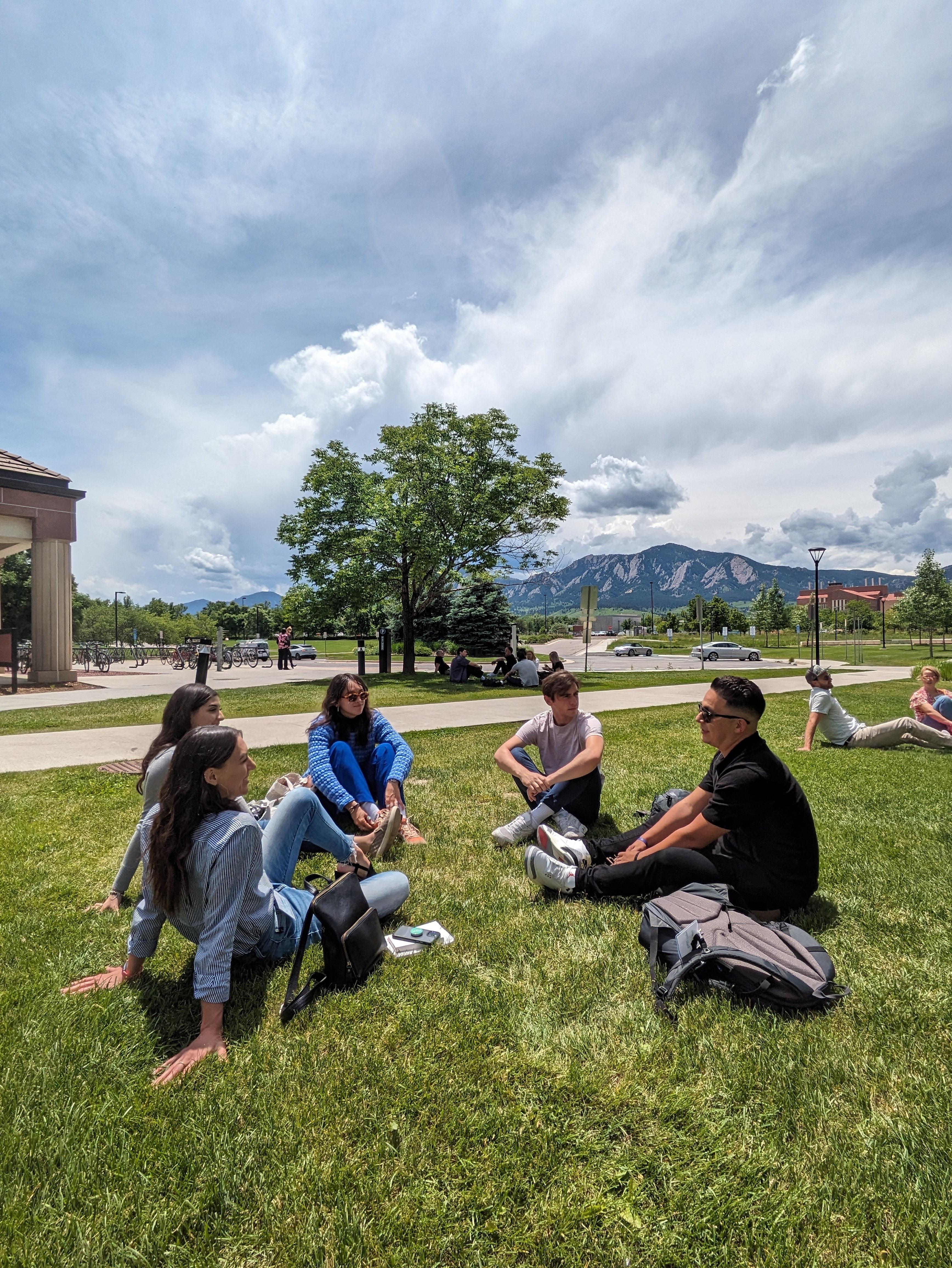 A group of young adults sitting on a grassy lawn under a bright sky with mountains and trees in the background, engaging in conversation and enjoying the outdoors.