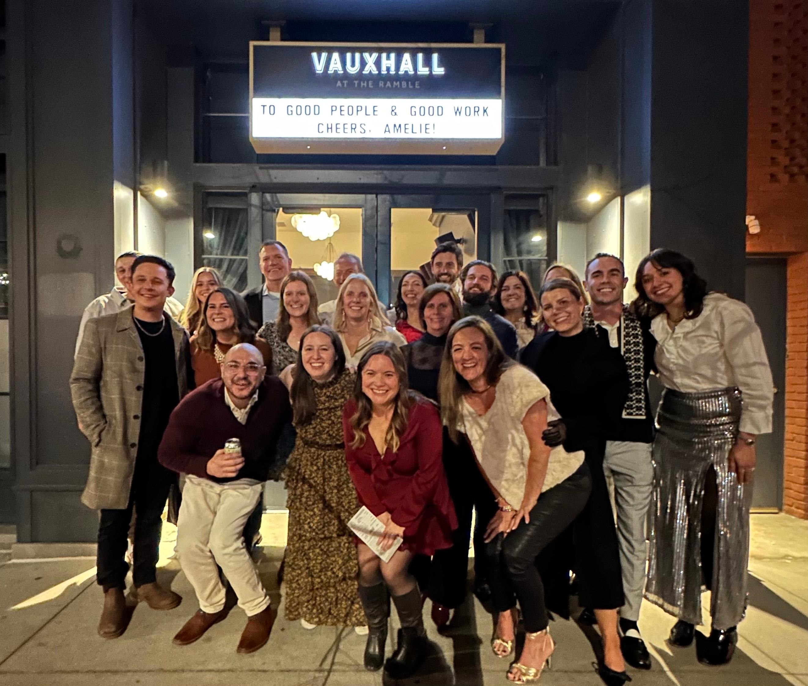 Group photo of smiling team members standing outside Vauxhall at The Ramble under a bright marquee that reads, 'To good people & good work. Cheers, Amelie!