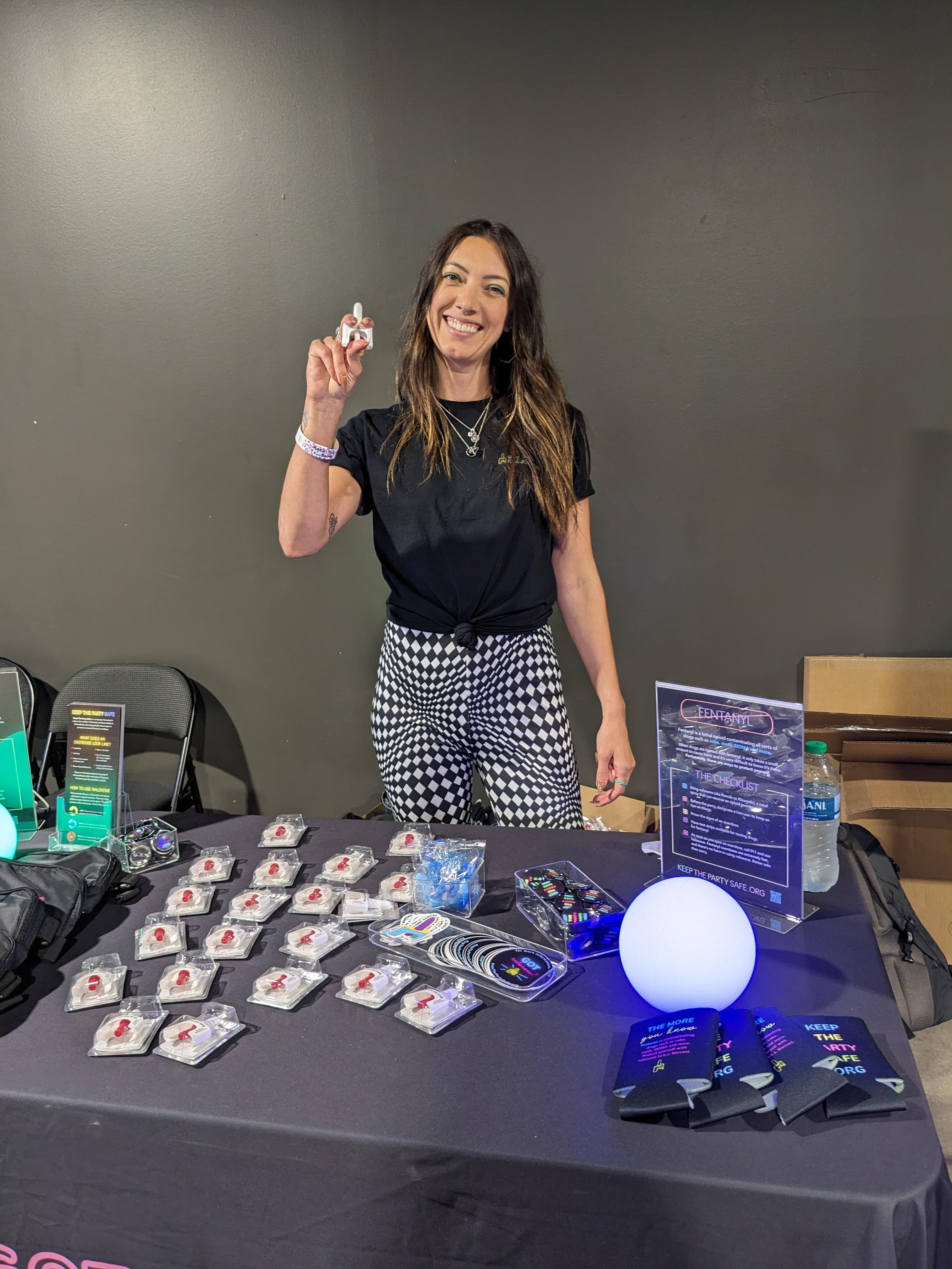 Smiling woman standing behind a booth displaying educational materials, promotional items, and merchandise for a 'Keep the Party Safe' campaign.