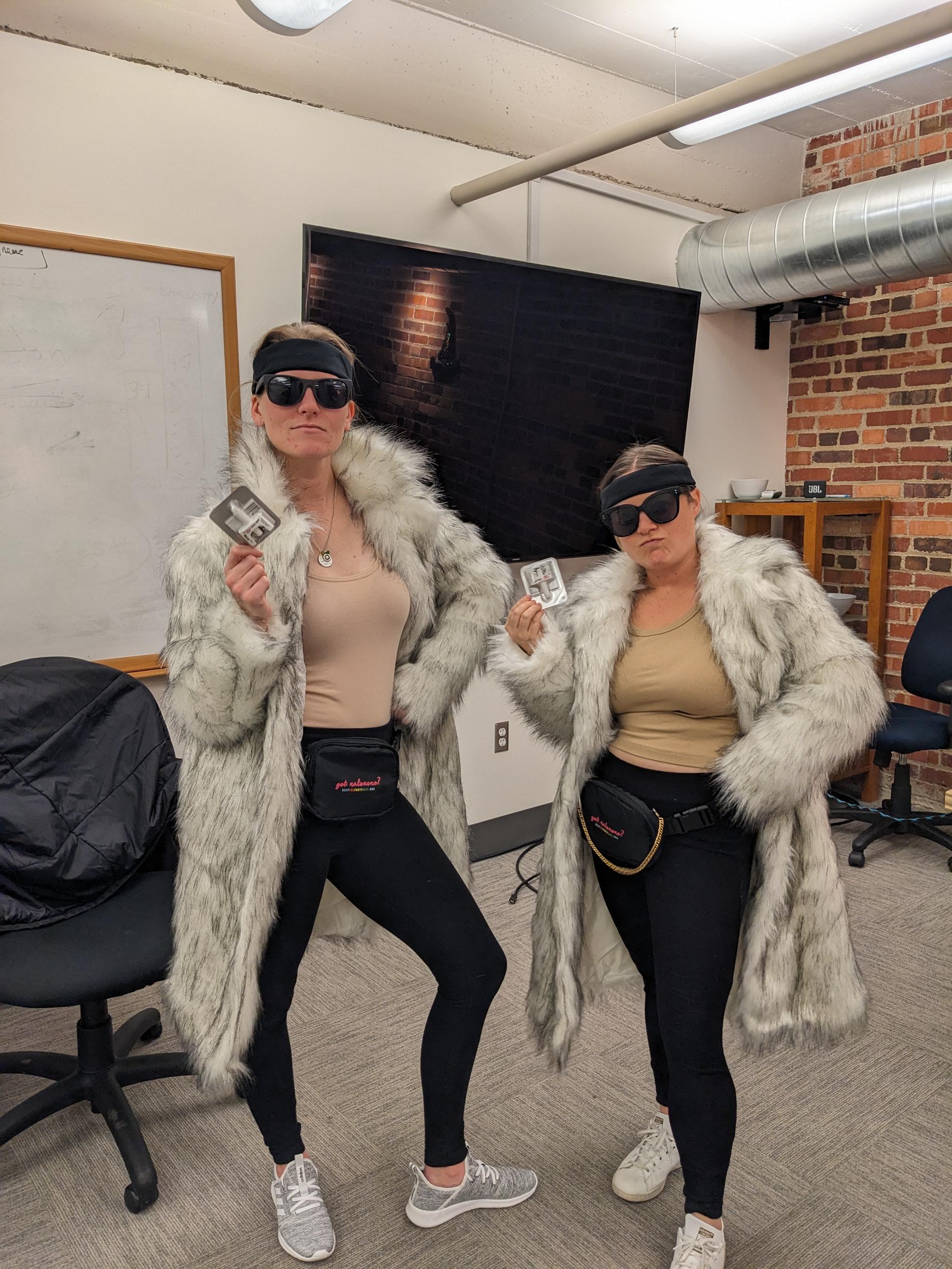 Two women dressed in matching faux fur coats, black leggings, and sunglasses, holding square promotional cards and posing confidently in an office setting.