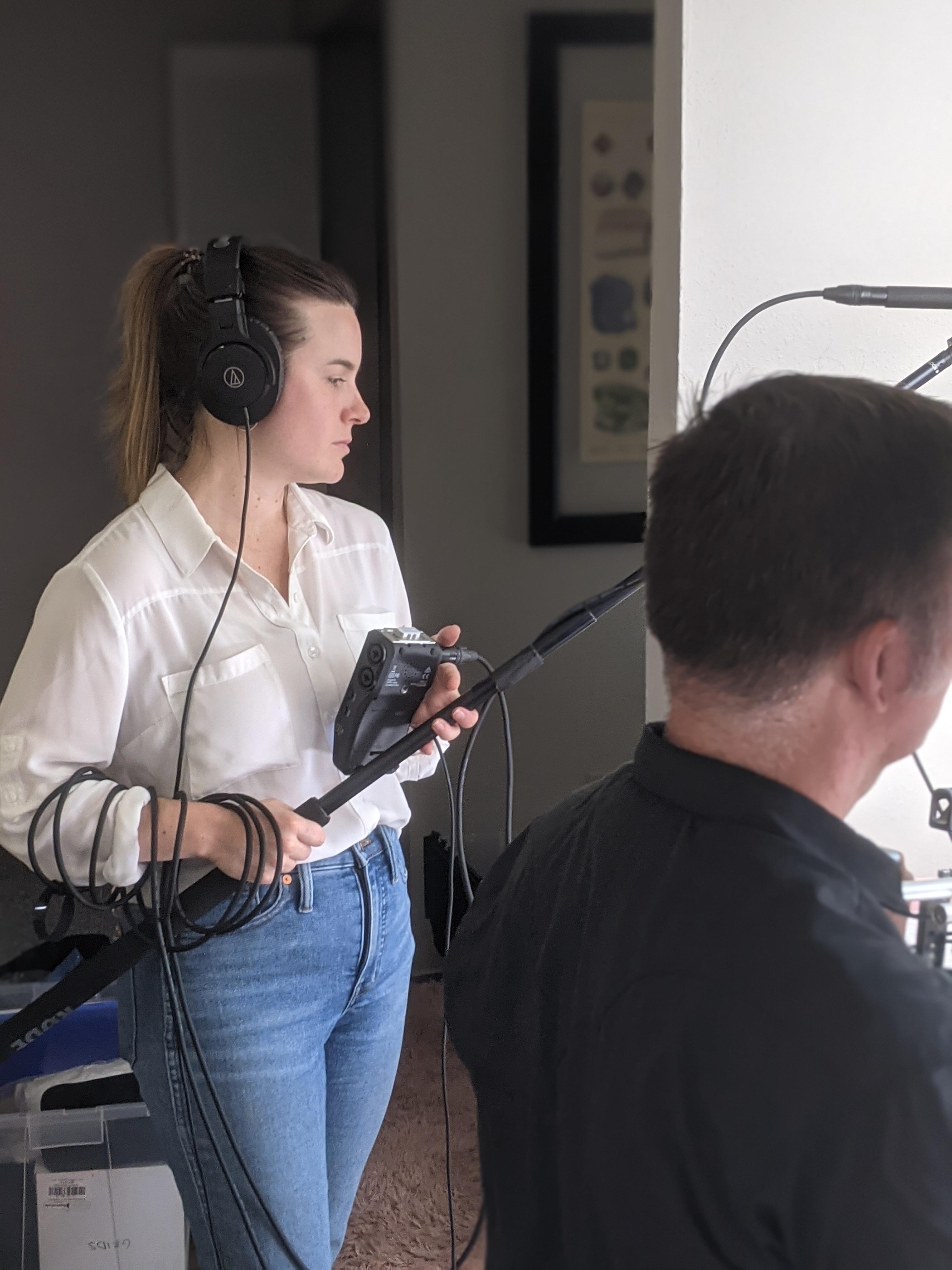 A woman wearing headphones and holding audio recording equipment while assisting in a video production setup, with another person in the foreground.