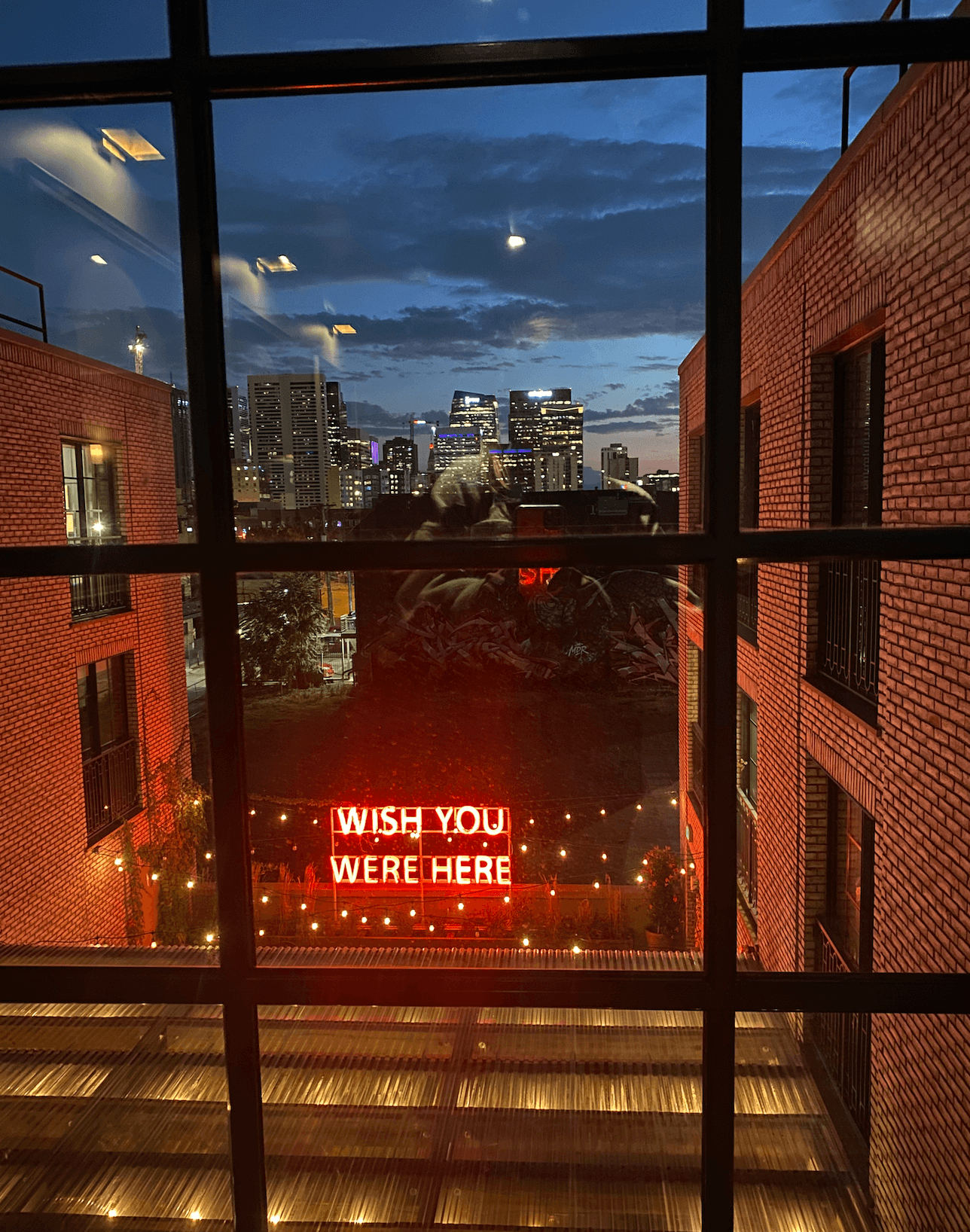 View through a window of a cityscape at dusk with illuminated buildings in the background and a neon sign reading 'Wish You Were Here' surrounded by string lights in the foreground.