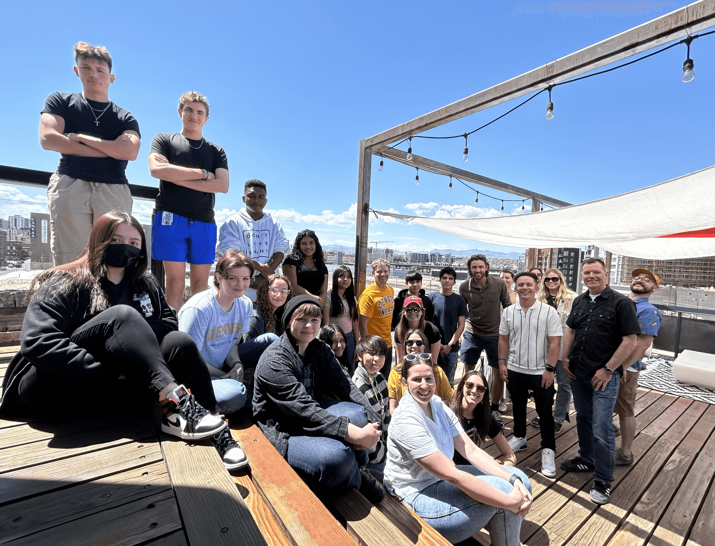 A group of people posing together on a rooftop deck under a sunny blue sky, with a cityscape in the background.