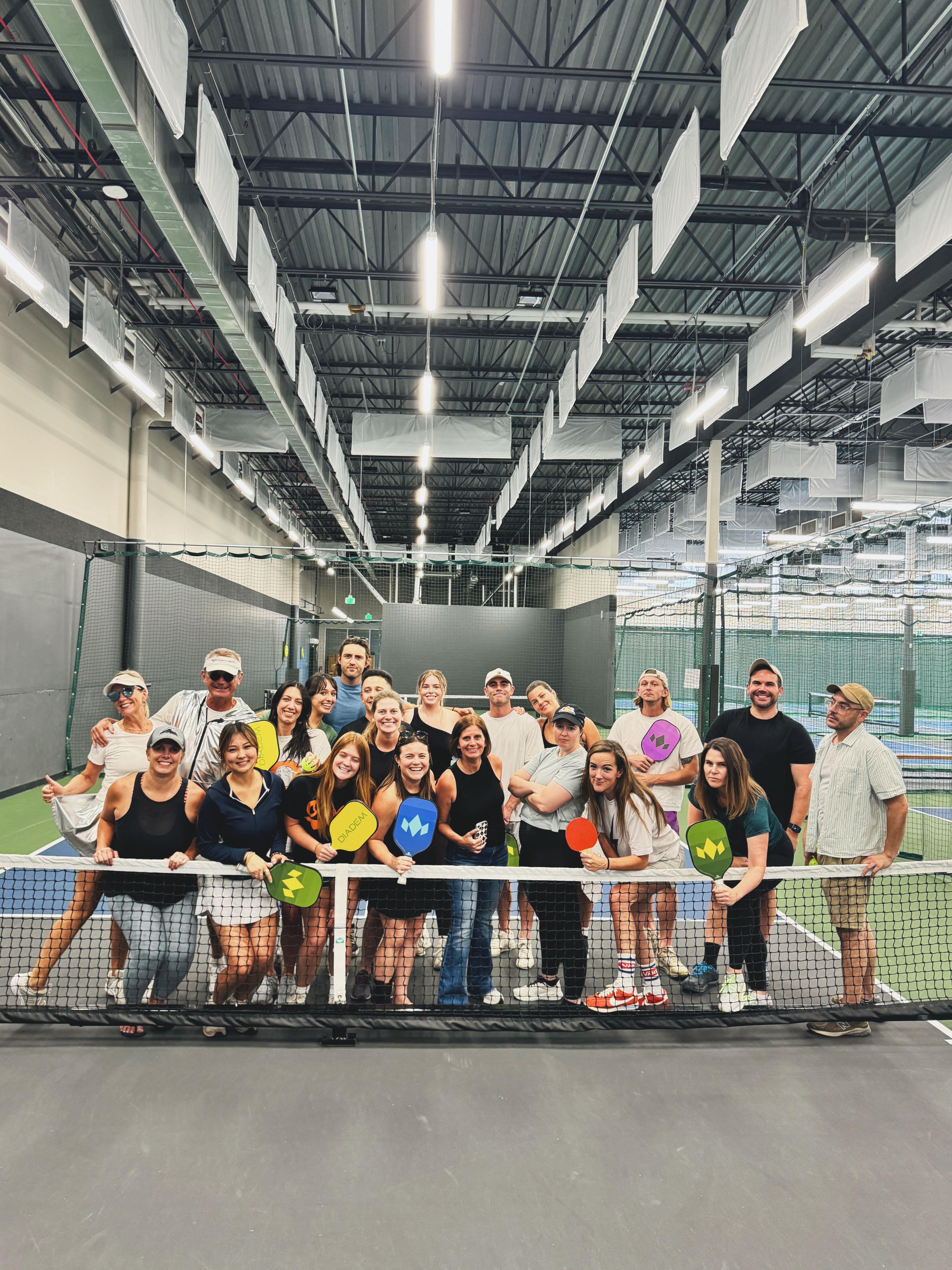 A group of people holding colorful paddles, standing on an indoor pickle ball court, smiling in front of a net under bright industrial lighting.