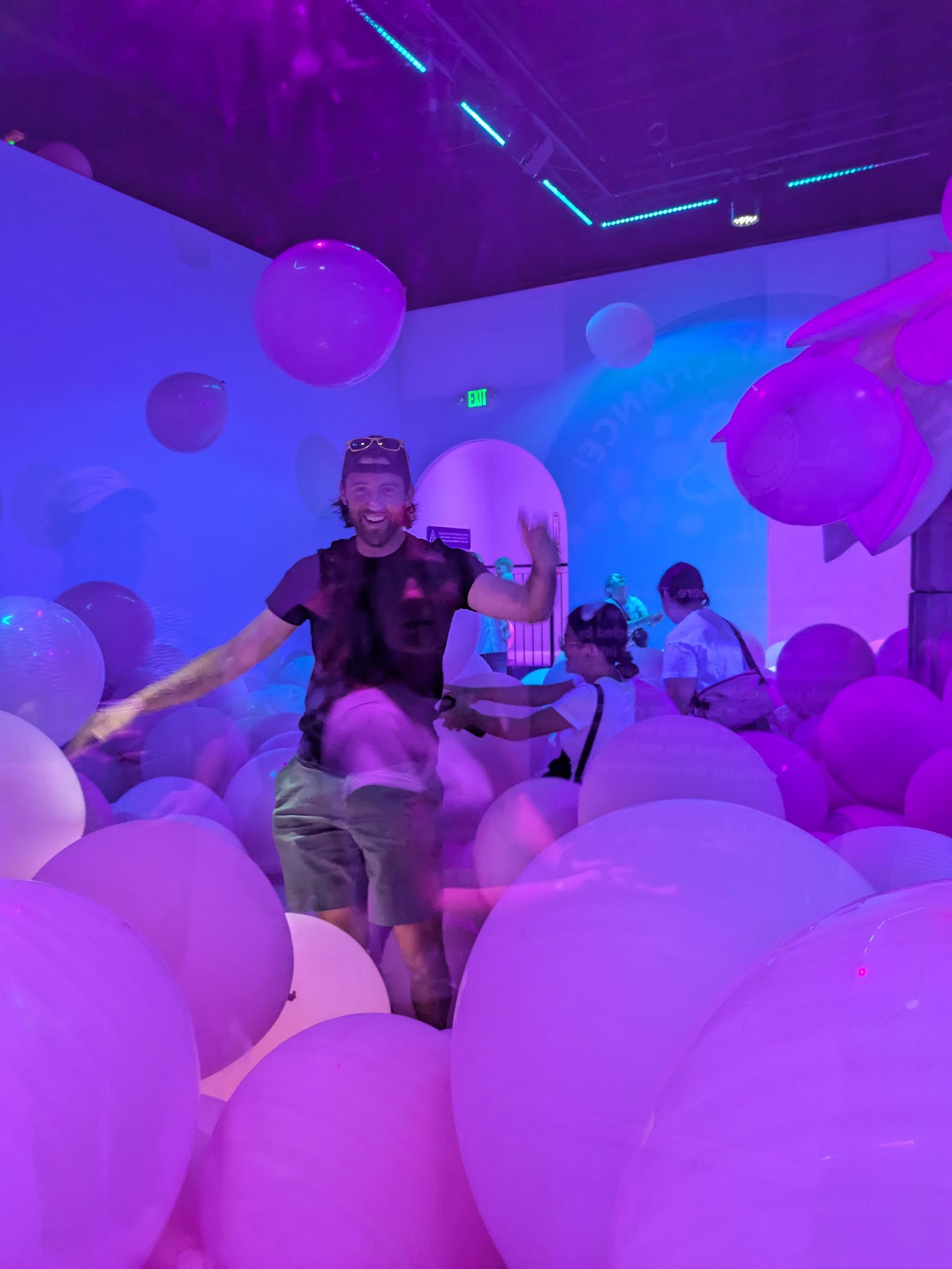 A man smiling and playing in a room filled with large pink and purple balloons under vibrant neon lighting, creating a playful and immersive atmosphere.