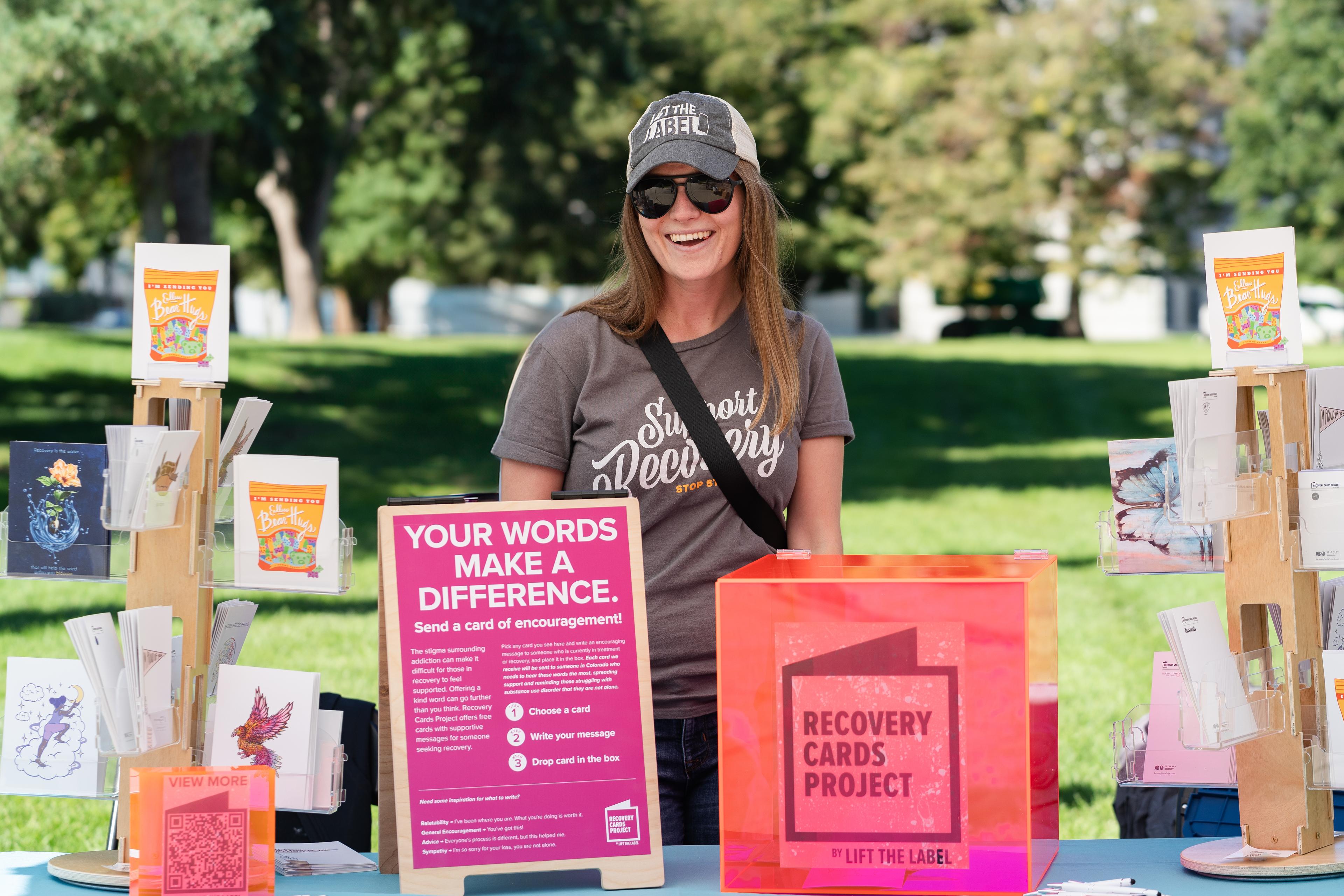 A woman wearing a 'Support Recovery' t-shirt and sunglasses standing at a booth for the Recovery Cards Project, featuring cards, a bright orange collection box, and a sign encouraging messages of support.